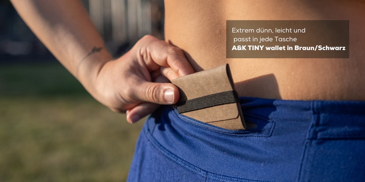 Ein A&K TINY Wallet wird von einer Frau mühelos in ihre Sporthosentasche gesteckt. Diese Darstellung verdeutlicht, wie klein und leicht das Wallet ist und wie problemlos es in jede Tasche passt.